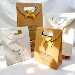 May include: Four gift bags in shades of brown and white, each with a handle and a decorative bow. The bags feature the words "Thank You" and names with a date. The bags are suitable for weddings or other celebrations.