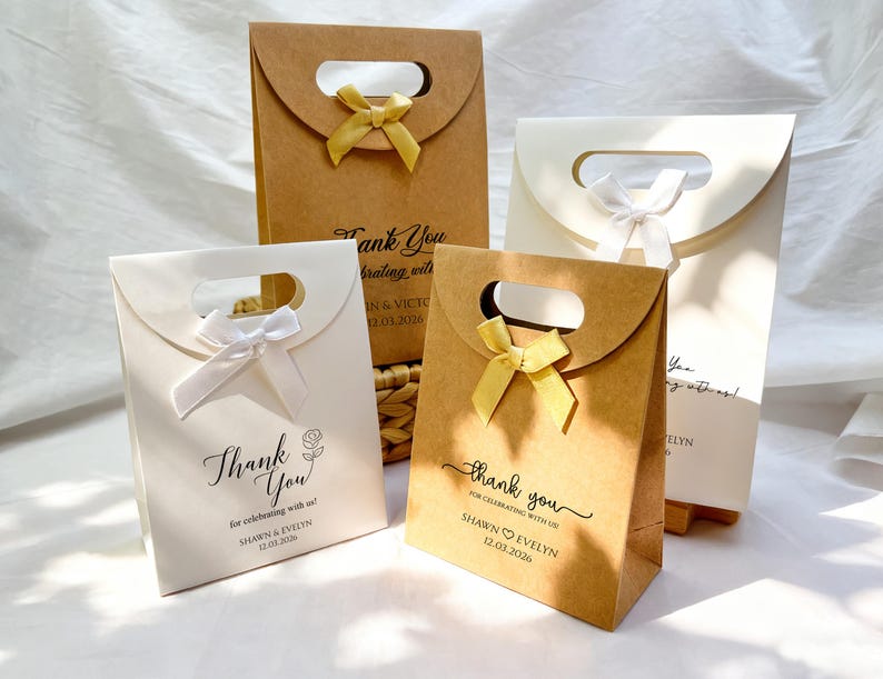 May include: Four gift bags in shades of brown and white, each with a handle and a decorative bow. The bags feature the words "Thank You" and names with a date. The bags are suitable for weddings or other celebrations.