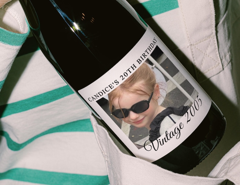 May include: A dark wine bottle with a label featuring a photo of a young girl wearing sunglasses. The label reads "CANDICE'S 20TH BIRTHDAY" and "Vintage 2005". The bottle is held against a striped shirt.