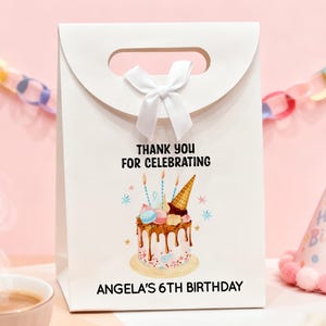 May include: White gift bag with a handle and a white bow. The bag features a birthday cake illustration with the text "THANK YOU FOR CELEBRATING" and "ANGELA'S 6TH BIRTHDAY". A party hat is visible in the background.