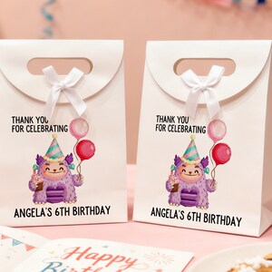 May include: Two white gift bags with a handle and a white bow. Each bag features a cartoon monster holding balloons and a cupcake. Text on the bags reads "THANK YOU FOR CELEBRATING" and "ANGELA'S 6TH BIRTHDAY".