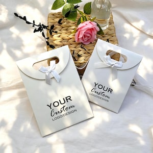 May include: Two white gift bags with a handle and a small white bow. The bags have the text "YOUR Custom LOGO/DESIGN" printed in black. A pink rose and greenery are in the background.