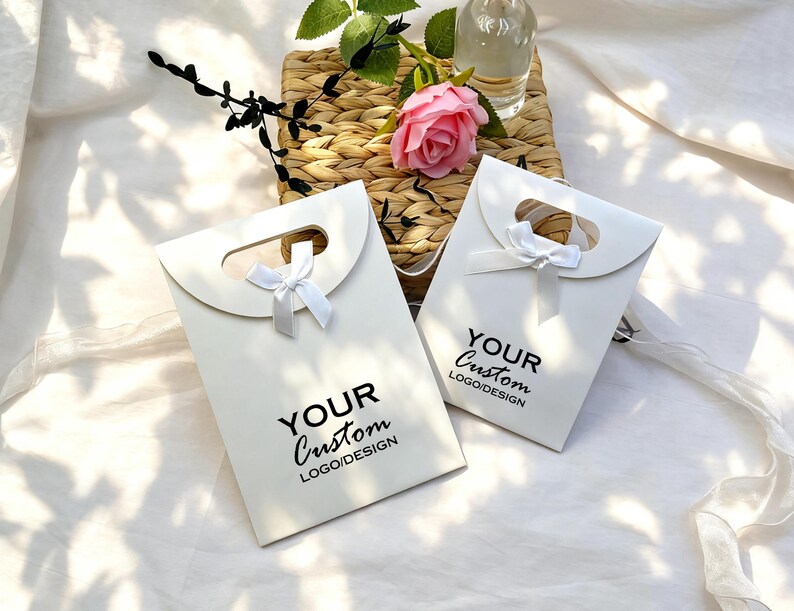 May include: Two white gift bags with a handle and a small white bow. The bags have the text "YOUR Custom LOGO/DESIGN" printed in black. A pink rose and greenery are in the background.