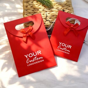 May include: Two red gift bags with a handle and a small red bow. The bags have the text "YOUR Custom LOGO/DESIGN" printed in white. The bags are on a white surface with a woven basket in the background.