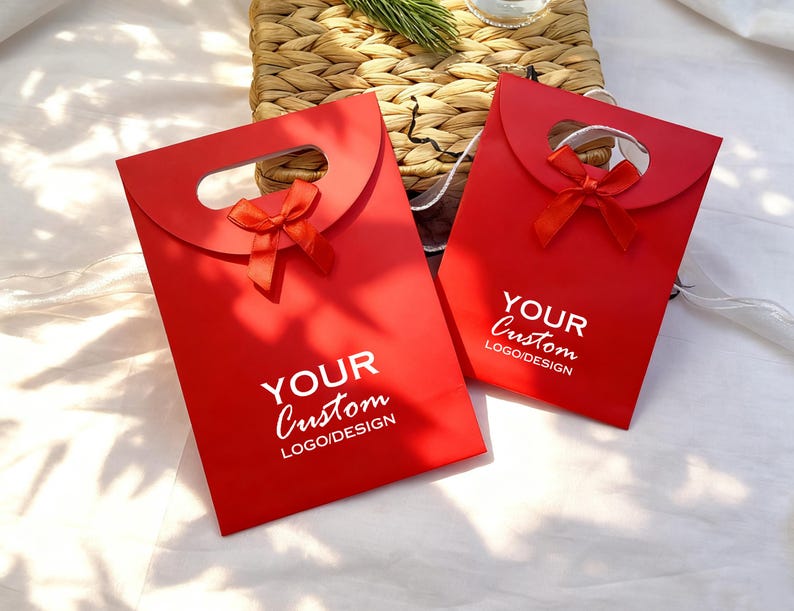 May include: Two red gift bags with a handle and a small red bow. The bags have the text "YOUR Custom LOGO/DESIGN" printed in white. The bags are on a white surface with a woven basket in the background.