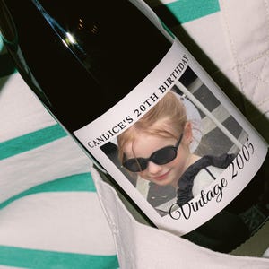 May include: A dark wine bottle with a label featuring a photo of a young girl wearing sunglasses. The label reads "CANDICE'S 20TH BIRTHDAY" and "Vintage 2005". The bottle is held against a striped shirt.