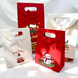 May include: Four Christmas gift bags in red and white. Each has a handle and a bow. The red bags feature a snowman and a Christmas elf graphic. The white bags have a gnome and gift graphic. Text reads "Merry Christmas."