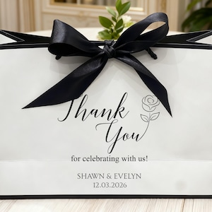 May include: A white gift bag with black trim and a large black satin bow. The bag features the words "Thank You" in elegant script, along with a rose illustration and the text "for celebrating with us!" and names and a date.