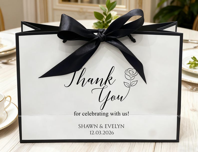May include: A white gift bag with black trim and a large black satin bow. The bag features the words "Thank You" in elegant script, along with a rose illustration and the text "for celebrating with us!" and names and a date.