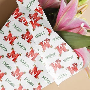 May include: White wrapping paper featuring a repeating pattern of a red letter "M" with Christmas decorations and the name "Melissa" in green. The paper is wrapped around a bouquet of pink lilies.