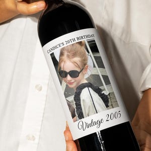 May include: A black wine bottle with a white label featuring a photo of a young girl wearing sunglasses. The label reads "CANDICE'S 20TH BIRTHDAY" and "Vintage 2005". The bottle is held by a person wearing a white shirt.