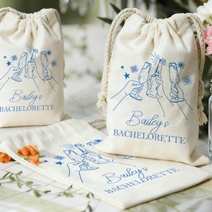 Könnte beinhalten: Cremefarbene Kordelzugbeutel mit blauen Illustrationen und Text. Das Design zeigt Hände, die auf Sektgläser und eine Flasche anstoßen, mit den Worten "Bailey's BACHELORETTE". Die Taschen liegen auf einem Tisch mit Blumen.