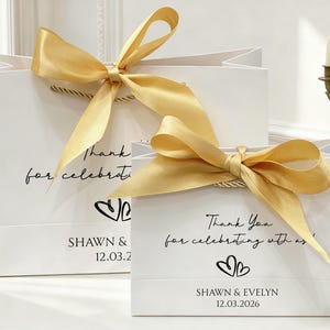 May include: Two white gift bags with gold satin bows. The bags have a handwritten font that says "Thank you for celebrating with us!" and the names "SHAWN & EVELYN" with the date 12.03.2026.