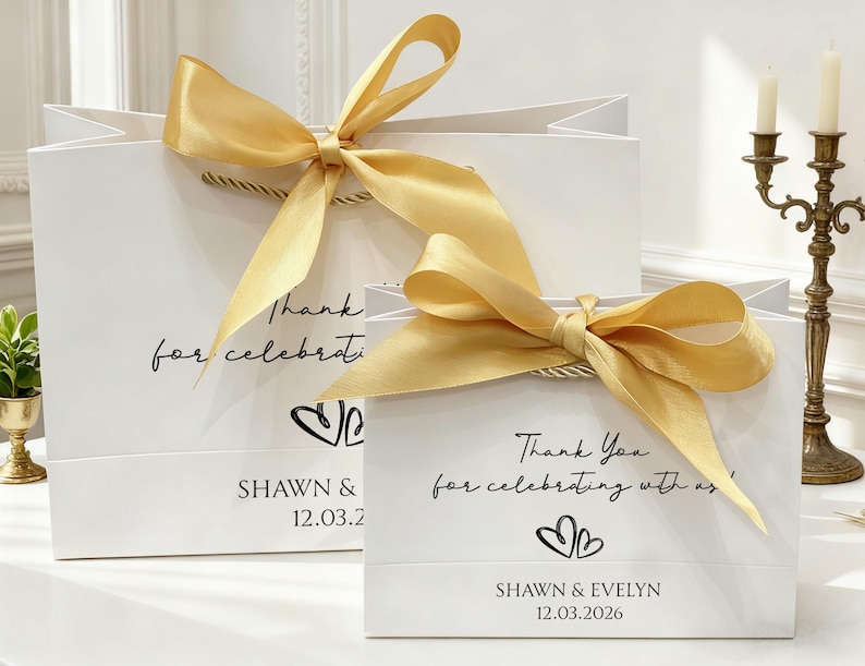 May include: Two white gift bags with gold satin bows. The bags have a handwritten font that says "Thank you for celebrating with us!" and the names "SHAWN & EVELYN" with the date 12.03.2026.