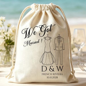 Peut inclure: Un sac à cordon en tissu couleur crème avec le texte imprimé en noir We Got Married! accompagné d'illustrations d'une robe de mariée et d'un costume. Sous les graphiques, les lettres D & W sont imprimées au-dessus de French Riviera 10.10.2028.