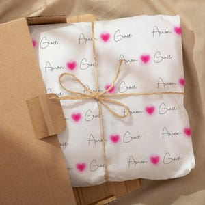 May include: A wrapped package in a cardboard box. The white wrapping paper is printed with the words "Grace" and "Apron" in a script font, along with pink heart shapes. The package is tied with twine.