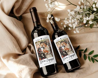 May include: Two dark glass wine bottles with custom labels. The labels feature a photo of a couple and the text "Karen Shawn We're Engaged 09.23.2026". The bottles are displayed on a beige fabric with white flowers.
