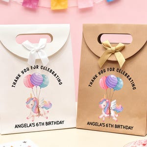 May include: Two gift bags, one white and one brown, with a handle and a bow. Each bag features a unicorn with balloons and the text "Thank you for celebrating" and "Angela's 6th Birthday."