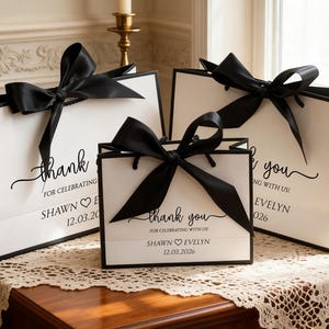 May include: Three white gift bags with black trim and large black satin bows. The bags have the words "thank you" and "for celebrating" printed on them, along with names and a date. The bags are on a wooden table.