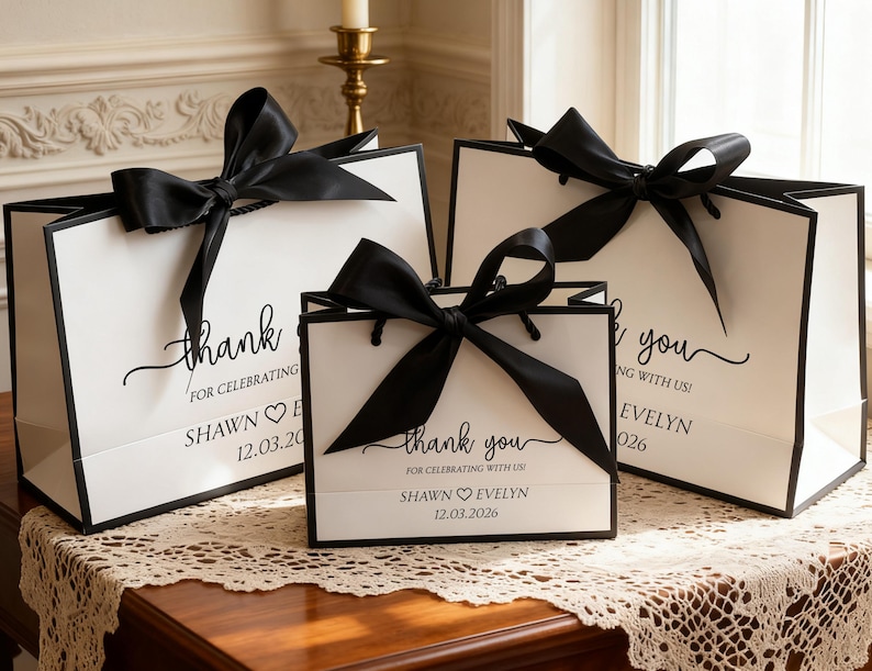 May include: Three white gift bags with black trim and large black satin bows. The bags have the words "thank you" and "for celebrating" printed on them, along with names and a date. The bags are on a wooden table.