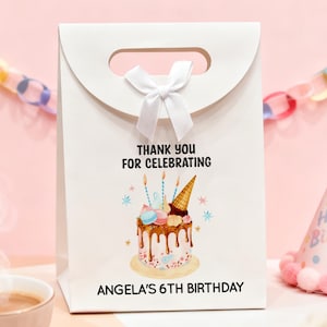May include: White gift bag with a handle and a white bow. The bag features a birthday cake illustration with the text "THANK YOU FOR CELEBRATING" and "ANGELA'S 6TH BIRTHDAY". A party hat is visible in the background.