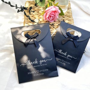 May include: Two black gift bags with a handle cutout and a black bow. The bags have the text "thank you" in cursive, along with the names "Shawn Evelyn" and the date "12.03.2026". A pink rose and greenery are in the background.
