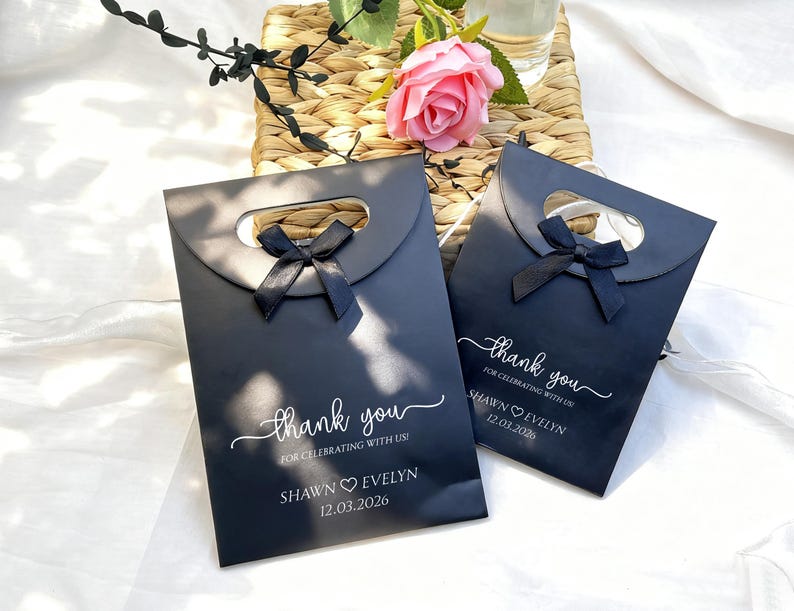 May include: Two black gift bags with a handle cutout and a black bow. The bags have the text "thank you" in cursive, along with the names "Shawn Evelyn" and the date "12.03.2026". A pink rose and greenery are in the background.