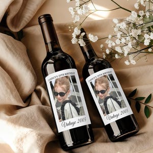 May include: Two dark glass wine bottles with custom labels. The labels feature a photo of a young person wearing sunglasses, with the text "Candice's 20th Birthday" and "Vintage 2005". The bottles are on a beige surface.
