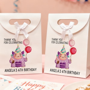 May include: Two white gift bags with a handle and a white bow. Each bag features a cartoon monster holding balloons and a cupcake. Text on the bags reads "THANK YOU FOR CELEBRATING" and "ANGELA'S 6TH BIRTHDAY".