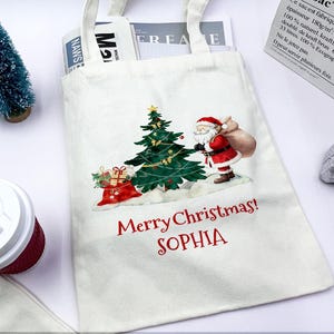 May include: White canvas tote bag with a Christmas design featuring Santa Claus, a decorated tree, and the text "Merry Christmas! SOPHIA" in red. The bag has white handles and is filled with magazines.