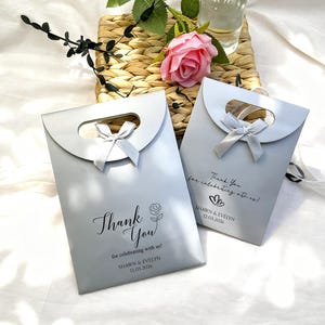 May include: Two silver gift bags with a curved handle and a bow. The bags have the text "Thank You" and "for celebrating with us!" printed on them, along with names and a date. A pink rose and greenery are in the background.