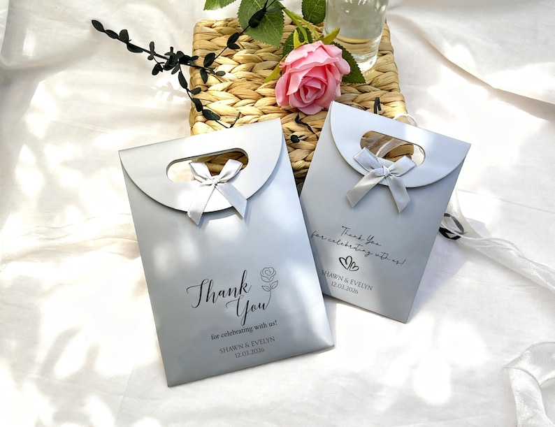 May include: Two silver gift bags with a curved handle and a bow. The bags have the text "Thank You" and "for celebrating with us!" printed on them, along with names and a date. A pink rose and greenery are in the background.