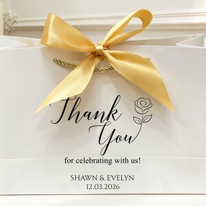 May include: White gift bag with a gold satin ribbon bow and rope handles. The bag features the words "Thank You" in elegant script, along with a rose illustration and the text "for celebrating with us!" and the names "SHAWN & EVELYN" and the date "12.03.2026".