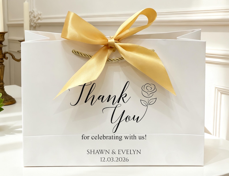 May include: White gift bag with a gold satin ribbon bow and rope handles. The bag features the words "Thank You" in elegant script, along with a rose illustration and the text "for celebrating with us!" and the names "SHAWN & EVELYN" and the date "12.03.2026".