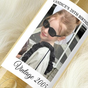 May include: A bottle with a label featuring a photo of a person wearing sunglasses. The label reads "Candice's 20th Birthday" and "Vintage 2005" in a script font. The bottle is on a white, fluffy surface.