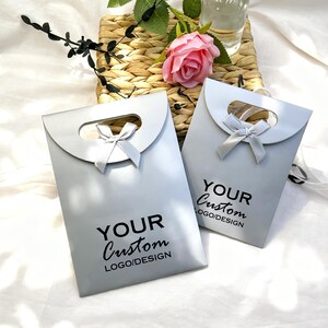 May include: Two light gray gift bags with a curved handle and a small bow. The bags have the text "YOUR Custom LOGO/DESIGN" printed in black. The bags are on a white surface with a woven basket and a pink rose in the background.