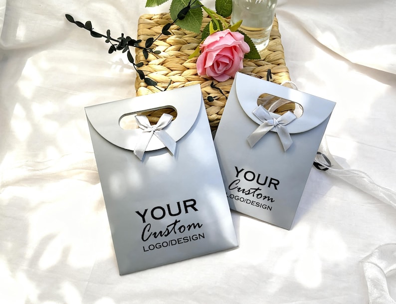 May include: Two light gray gift bags with a curved handle and a small bow. The bags have the text "YOUR Custom LOGO/DESIGN" printed in black. The bags are on a white surface with a woven basket and a pink rose in the background.