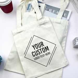 May include: Two off-white canvas tote bags with two handles each. The front bag features a black diamond-shaped graphic outline containing the text "YOUR CUSTOM LOGO/TEXT HERE" in a bold, sans-serif font centred within the geometric frame.