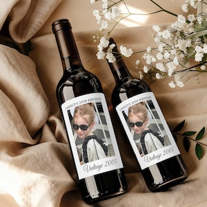 May include: Two dark glass wine bottles with custom labels. The labels feature a photo of a young person wearing sunglasses, with the text "Candice's 20th Birthday" and "Vintage 2005". The bottles are displayed on a beige surface.
