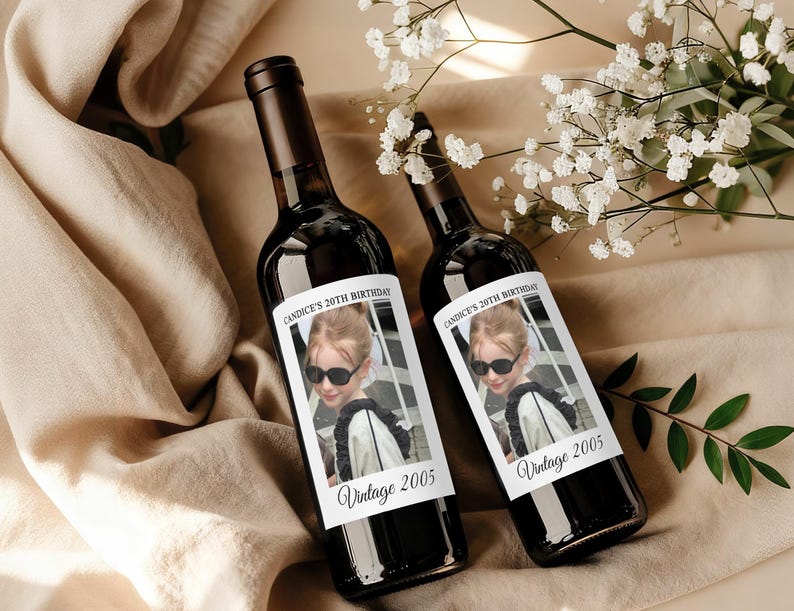 May include: Two dark glass wine bottles with custom labels. The labels feature a photo of a young person wearing sunglasses, with the text "Candice's 20th Birthday" and "Vintage 2005". The bottles are displayed on a beige surface.