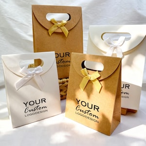 May include: Four gift bags in the image. Two are kraft paper, and two are white. Each bag has a handle and a ribbon bow. The text "YOUR Custom LOGO/DESIGN" is printed on two of the bags. The other two bags have the text "DUR stom DIDESIGN" and "JR om SIGN".