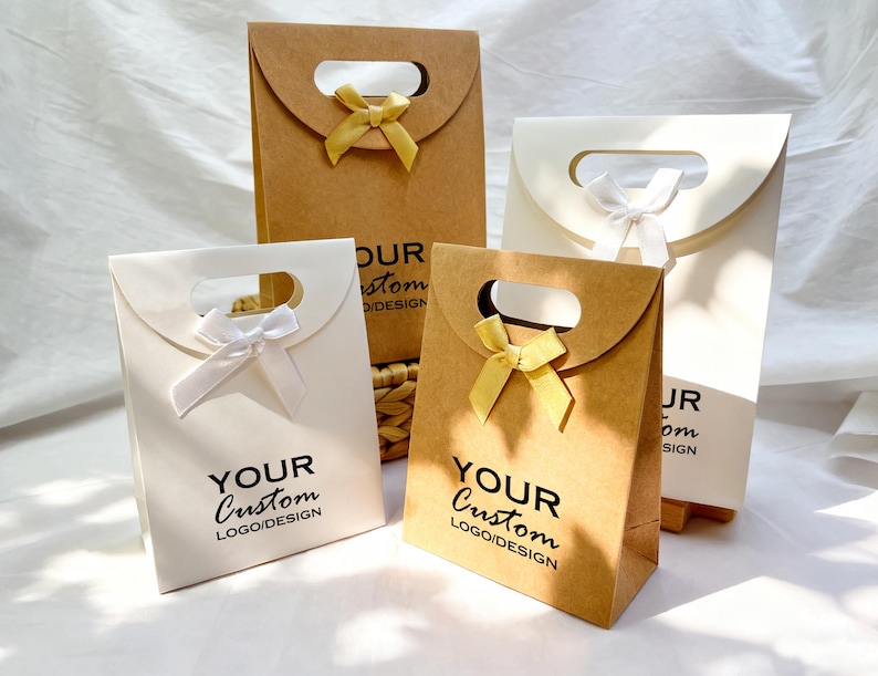 May include: Four gift bags in the image. Two are kraft paper, and two are white. Each bag has a handle and a ribbon bow. The text "YOUR Custom LOGO/DESIGN" is printed on two of the bags. The other two bags have the text "DUR stom DIDESIGN" and "JR om SIGN".