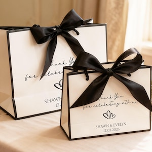 May include: Two white gift bags with black trim and large black satin bows. The bags have the text "Thank You for celebrating with us" and the names "Shawn & Evelyn" with a date. The bags are on a white surface.