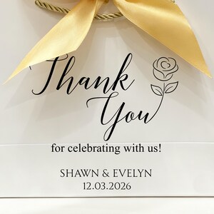 May include: White gift bag with a gold ribbon bow. The bag features the words "Thank You" in elegant script, a rose illustration, and the text "for celebrating with us!" along with names and a date.