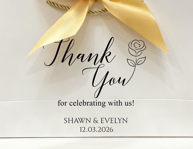 May include: White gift bag with a gold ribbon bow. The bag features the words "Thank You" in elegant script, a rose illustration, and the text "for celebrating with us!" along with names and a date.
