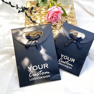 May include: Two black gift bags with a handle cutout and a black ribbon bow. The bags have the text "YOUR Custom LOGO/DESIGN" printed in white. A pink rose and greenery are in the background.