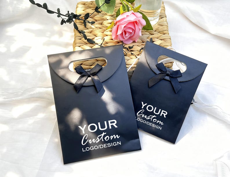 May include: Two black gift bags with a handle cutout and a black ribbon bow. The bags have the text "YOUR Custom LOGO/DESIGN" printed in white. A pink rose and greenery are in the background.