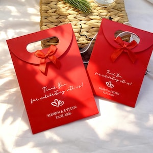 May include: Two red gift bags with a handle and a small red bow. The bags have the text "Thank You for celebrating with us!" and the names "SHAWN & EVELYN" with a date. The bags are on a white surface.