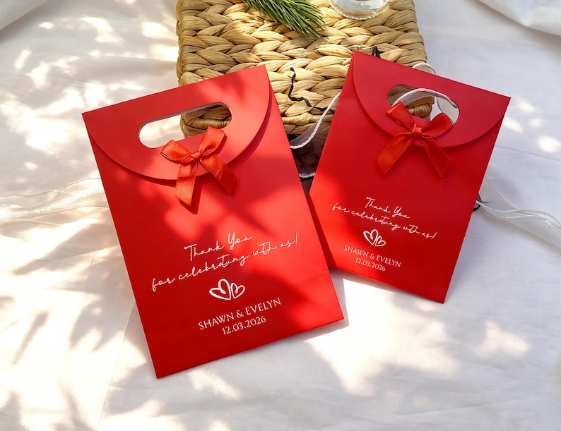May include: Two red gift bags with a handle and a small red bow. The bags have the text "Thank You for celebrating with us!" and the names "SHAWN & EVELYN" with a date. The bags are on a white surface.