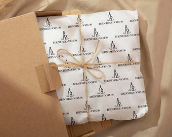 Puede incluir: Un paquete envuelto en papel blanco con un patrón repetido de botellas de vino y copas negras, atado con hilo, dentro de una caja de cartón marrón. El texto "RENDEZ-VOUS" está impreso en el papel de envolver.